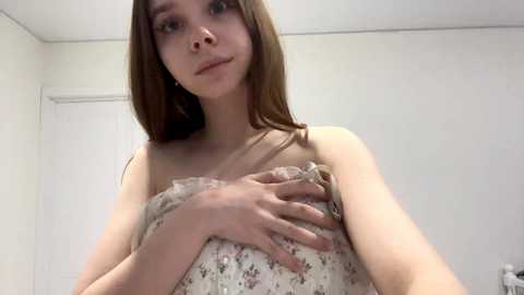 Video of a young Caucasian woman with long brown hair, wearing a floral-patterned, strapless dress, standing in a plain, white-walled room, with her right hand resting on her chest.