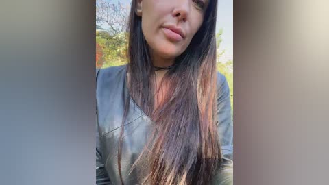 A video of a young woman with long, straight brown hair, wearing a black leather jacket and a choker necklace. She has a neutral expression and is standing outdoors, with blurred greenery and a blue sky in the background.