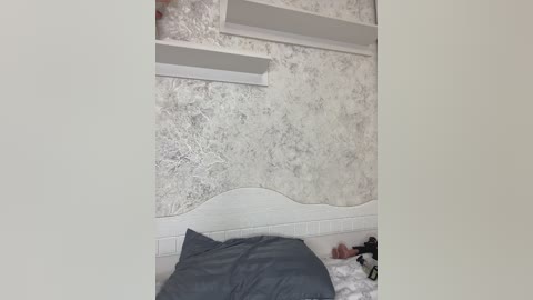 A video of a minimalist bedroom with a white headboard, gray bedding, and textured silver wallpaper with white shelves. The room has a modern, clean aesthetic with a focus on simplicity and functionality.