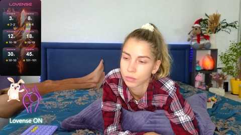 Video of a young woman with blonde hair in a ponytail, wearing a red plaid shirt, lying on a bed with a blue-patterned blanket, in a cozy bedroom setting.