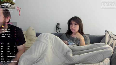 Video of a woman in a gray t-shirt, wrapped in a quilt, watching a man in a black shirt on a couch in a white room.
