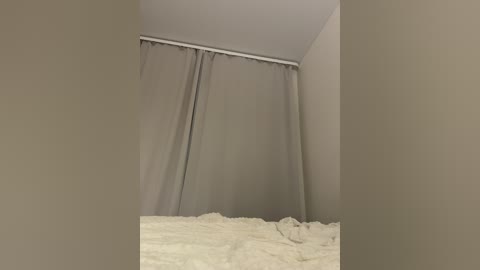 Video of a small, empty room with beige walls and a light brown curtain covering a window at the top. The bed, covered in a fluffy white comforter, occupies the bottom half of the image.