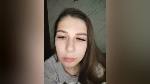 Video of a young woman with long brown hair, light skin, and a neutral expression, wearing a gray shirt. Background features a textured gray wall.