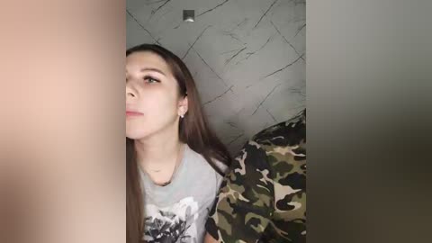 Video of a young woman with straight brown hair, wearing a camouflage-patterned shirt, looking upwards with a neutral expression. Background features textured gray wall and part of a camouflage-patterned pillow.