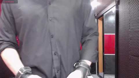 Video of a person wearing a black button-up shirt, white gloves, and black leather wrist restraints, standing in a dimly lit, modern hallway with red and black walls.