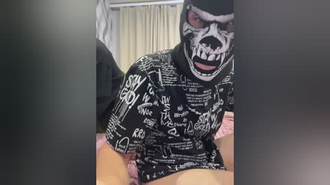 Video of a person wearing a black, white, and gray skeletal mask with an open mouth, accompanied by a black T-shirt with white scribbles. The background shows a beige curtain.