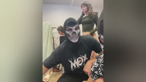 Video of a man in a black hoodie with a skull mask, sitting on a wooden bench, surrounded by two women in tight, revealing outfits, in a dimly lit, indoor setting.