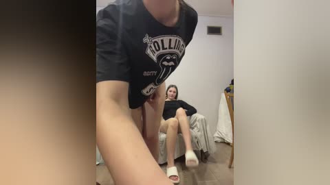 A video of a woman in a black \"Hollister\" t-shirt, revealing her bare thighs, and a woman in a black shirt and grey pants sitting on a bed, in a bedroom.