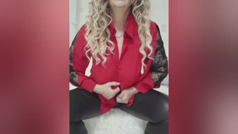 Video of a fair-skinned woman with long, wavy blonde hair, wearing a red lace shirt over black leggings, seated on a white surface.