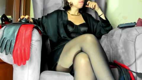 Video of a woman with medium skin tone, wearing black lingerie, sheer stockings, and a black robe, seated in a plush gray armchair, with red gloves draped over the armrest.