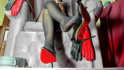 Video of a woman's legs in sheer black pantyhose and red-soled black stiletto heels, reclining on a plush grey sofa with a maroon throw blanket.