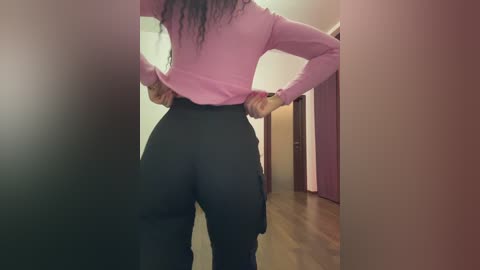 Video of a woman with long black hair in a pink shirt, pulling up black pants with a brown belt, in a hallway with wooden floors and purple doors.