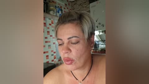 Video of a middle-aged woman with short, light grey hair, closed eyes, and slightly pursed lips, wearing a black necklace, in a rustic kitchen with red and white tiled walls and a whiteboard.