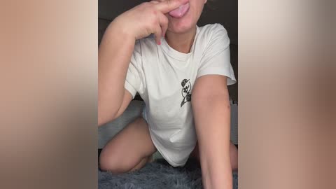 Video of a fair-skinned woman with a slender build, wearing a white t-shirt and no pants, kneeling on a plush gray carpet, holding her chin with her left hand.