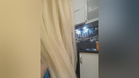 A video of a woman's blonde hair partially obscuring her face as she stands in a modern kitchen with white cabinets, a dark countertop, and a tiled backsplash featuring a cityscape mural.