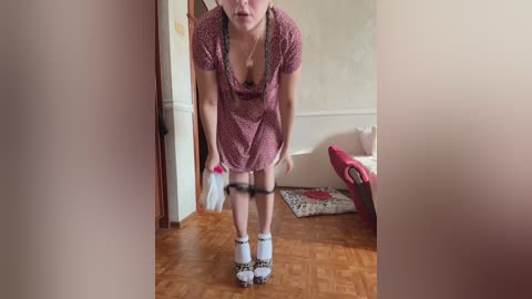 Video of a slender woman with fair skin, wearing a pink polka-dotted dress and white socks, bent over, pulling down black panties. Background: wooden floor, beige walls, red and white furniture.