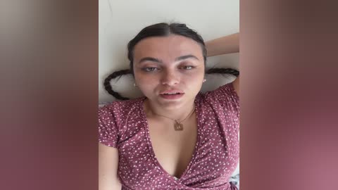 A video of a young woman with light brown skin, dark hair in braids, wearing a low-cut, pink floral dress, lying on a white bed, with blurred faces in the foreground.