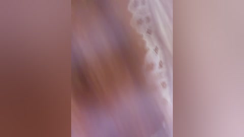 This image is a blurred video of a nude female figure with a light brown complexion. The focus is on her lower body, with a hint of a breast visible on the left side. The background is a neutral, beige wall, adding a soft, intimate atmosphere.