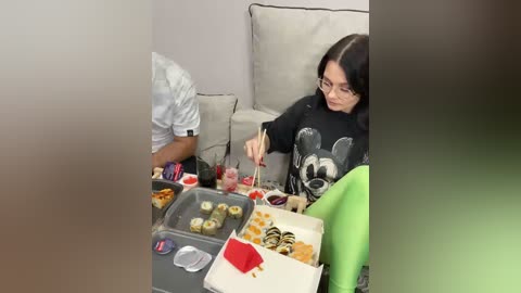 Video of a woman with glasses and black top, seated on a beige couch, eating sushi from a tray, surrounded by food, in a modern living room.