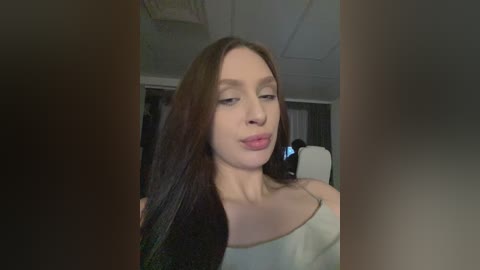 Video of a Caucasian woman with long brown hair, wearing a white off-shoulder top, puckering her lips in a playful manner. The background shows a dimly lit room with a white ceiling and a person in the distance.