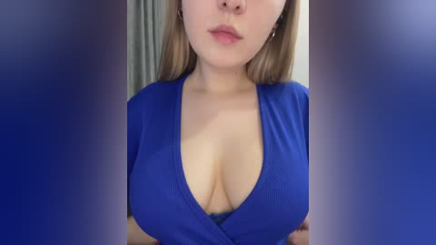 Video of a light-skinned woman with long, blonde hair, wearing a low-cut, deep blue dress that accentuates her large breasts. The background is blurred, possibly a bedroom setting.