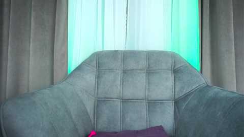 A video of a modern living room with a plush, light grey, tufted armchair in the foreground, flanked by beige curtains and a bright turquoise wall behind.