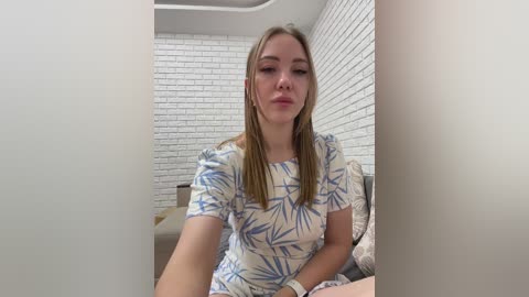 A video of a young woman with fair skin and long blonde hair, wearing a white dress with blue leaf patterns, sitting on a bed with a white brick wall background.