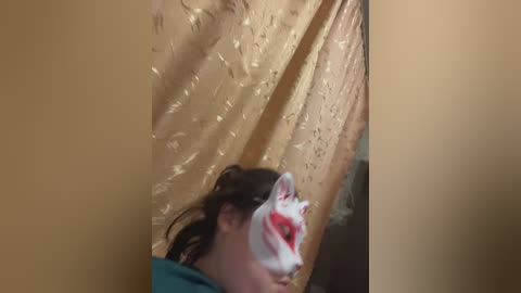 A video of a woman with dark hair and a white and red mask covering her face, standing in a dimly lit room with gold, patterned curtains.