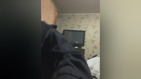Video of a person wearing a black jacket, standing in a small, cluttered room with a TV on a dresser, floral wallpaper, and a white bed.