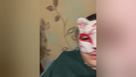 Video of a person wearing a white and red stylized fox mask with detailed markings, partially obscured by a teal garment, against a beige wall with a floral pattern.