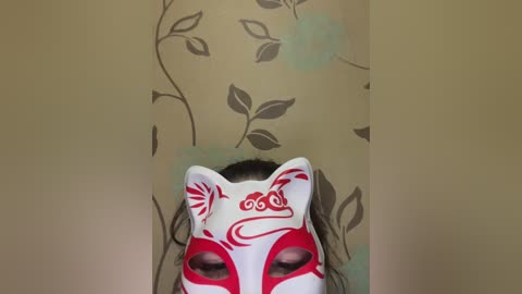 A video of a white fox mask with red and black swirling designs, placed against a beige wall adorned with brown floral and leaf patterns.