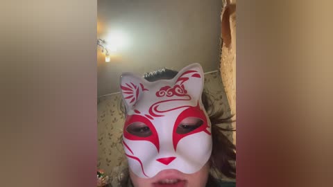 A video of a person lying on a bed, wearing a white fox mask with red markings and a black headband, in a dimly lit room with floral wallpaper.