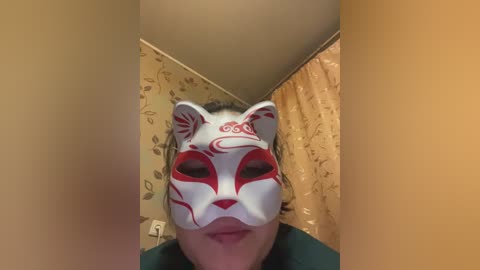Video of a person wearing a white fox mask with red designs, standing in a narrow hallway with beige walls and floral-patterned curtains.