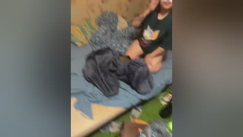 A blurry video shows a young person with light skin, wearing a dark green T-shirt and blue jeans, sitting on a bed. The bed has a blue blanket and a colorful pillow with yellow and green patterns. The room appears messy, with scattered items on the floor.