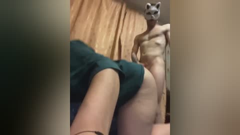 Video: A man in a bear mask, nude, stands in a room with beige curtains. A woman in teal shorts is on her knees in the foreground, legs visible.