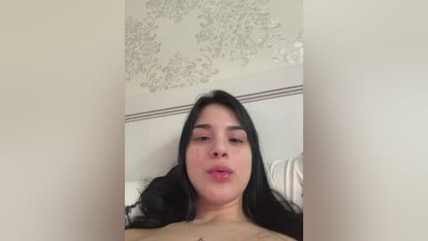 Video of a young woman with long black hair, light skin, and full lips, lying on a bed with a white headboard. The background shows an ornate, floral-patterned ceiling.