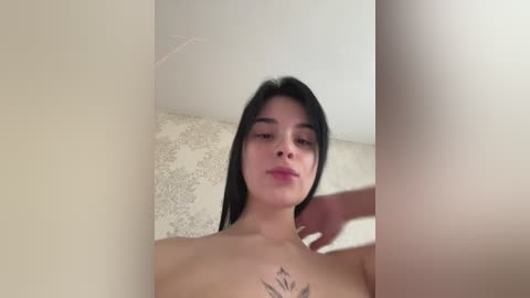 A video of a young woman with long black hair and light skin, topless, standing in a room with cream-colored wallpaper. She has a tattoo of a flower on her chest.