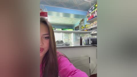 Video of a young woman with long brown hair, wearing a pink top, peering through a frosted glass door into a brightly lit, modern fast-food restaurant with a counter and shelves filled with food items.