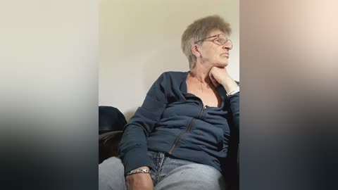 Video of an older woman with short gray hair, wearing glasses, a navy blue jacket, and jeans, sitting thoughtfully on a black couch, with a neutral beige wall in the background.