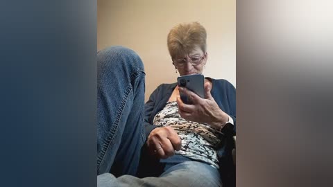 Video of an older woman with short, light-brown hair, wearing a patterned blouse and blue jeans, sitting and holding a smartphone, in a dimly lit room with beige walls.
