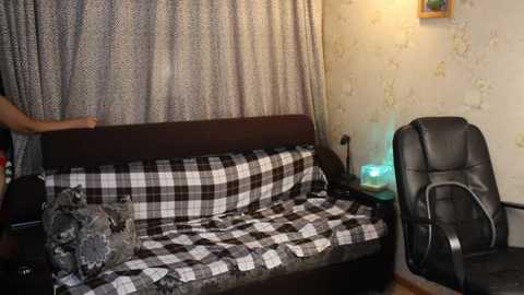 Video of a cozy, dimly-lit room with a black leather office chair, a plaid-patterned sofa, and beige wallpaper with floral designs.