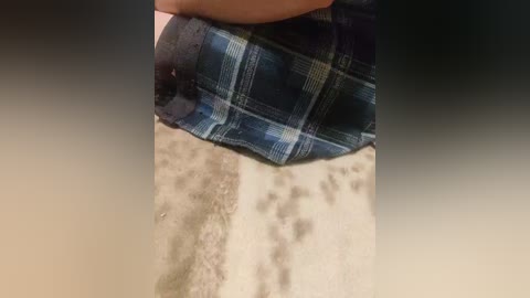Video of a person\u2019s hand pulling up a blue and white plaid skirt, revealing a sandy, uneven beach floor with footprints. The background is blurred, focusing on the intimate and casual scene.