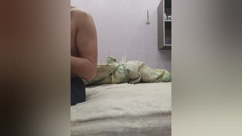 Video of a person lying on a bed with a green and white floral sheet, purple wall, and a black shelf with items.