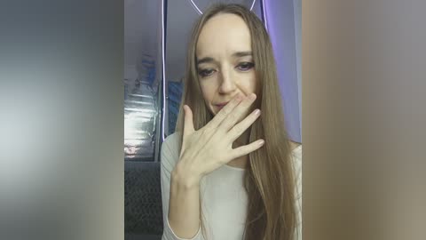 A video of a young woman with long brown hair, wearing a white shirt, covering her mouth with her right hand, standing in a dimly lit room with metallic walls and a partially visible window.