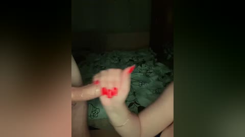 Video of a dimly lit bedroom featuring a man's hand with red nail polish gripping a woman's bare breast, both with fair skin. A green bedsheet with black cartoon characters is visible in the background.