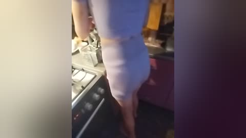 Video of a woman in a tight, light blue dress, standing in a dimly lit kitchen with a stove and cabinets in the background.
