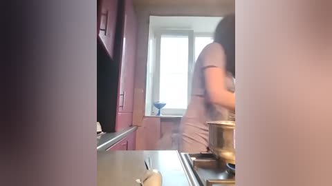 Video of a woman in a tight, pink dress stirring a pot on a stove in a small, dimly lit kitchen with pink cabinets and a window.