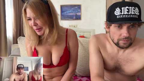 Video of a blonde woman in red lingerie and a man in a black cap, both in a bedroom setting.