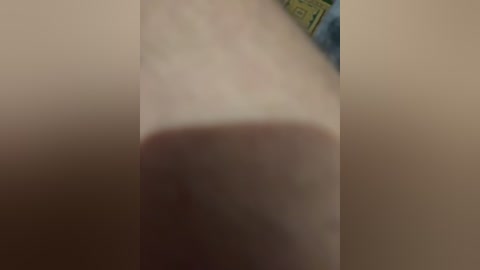 A close-up video of a large, erect penis, with skin tone appearing pale and slightly reddened at the tip, positioned centrally in the frame. The background is blurred, featuring a green and yellow patterned object to the right.