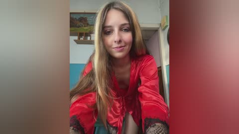 Video of a young woman with long, straight blonde hair, wearing a red satin robe with lace trim, standing in a room with white walls and a framed painting in the background.